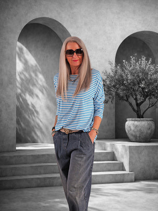 Frau im OFF#DLY Ringelshirt Frida türkis/white, gestreiftes Longsleeve mit lockerem Schnitt. Sie trägt es mit dunklen Jeans und Accessoires, vor modernem, architektonischem Hintergrund stehend.