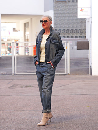 Frau trägt OFF#DLY Denim Jogger darkwash, dunkel gewaschen, locker geschnitten, hochgekrempelt, kombiniert mit passender Jacke und beigefarbenen Stiefeletten, Hände in den Taschen, auf Stadtstraße.