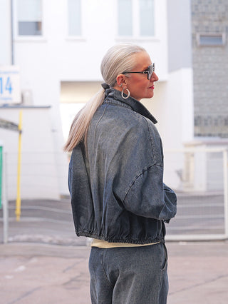 Person trägt die OFF#DLY Denim Jacke Ornella darkwash mit elastischem Saum und großer Brusttasche, kombiniert mit passenden Denim-Pants. Sie steht vor moderner Gebäude-Fassade und schaut nach rechts.