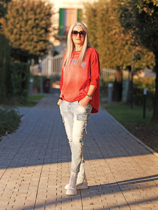 Ältere Frau trägt OFF#DLY Denim Verona blue: legere Cargo-Jeans mit Taschen und leichten Used-Elementen, kombiniert mit rotem Sweatshirt und weißen Sneakers, in einem baumgesäumten Park bei Sonnenuntergang.