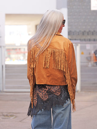 Person von hinten in urbaner Umgebung, trägt die OFF#DLY Jacke Rodeo camel, eine karamellfarbene, kurz geschnittene Jacke mit langen Fransen. Darunter ein Spitzen-Top, dazu locker geschnittene Jeans.