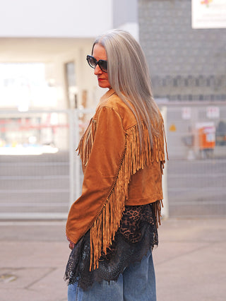 Person trägt die OFF#DLY Jacke Rodeo camel, eine Veloursjacke mit auffälligen Fransen an Rücken und Ärmeln, kombiniert mit einem Spitzenoberteil und Jeans in urbaner Umgebung.