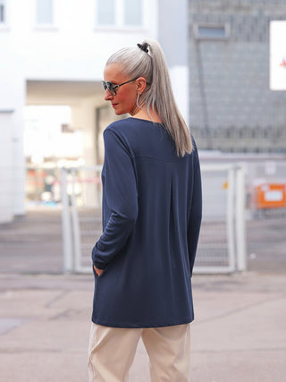 Frau im urbanen Umfeld trägt das OFF#DLY Shirt Eva darkblue, ein langärmeliges, navyfarbenes Oberteil mit Taschen und Rundhalsausschnitt. Kombiniert mit hellen Hosen und Sonnenbrille.