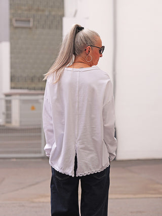Person von hinten in urbanem Umfeld, trägt das OFF#DLY LS Fiona white Langarmshirt mit Spitzensaum und Rückenschlitz, kombiniert mit dunkler Hose. Silberner Pferdeschwanz und Sonnenbrille ergänzen den Look.