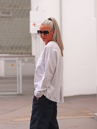 Eine Person in einem weißen OFF#DLY LS Fiona Longsleeve mit Spitzensaum, entspanntem Schnitt und verlängertem Rücken, steht lässig draußen.