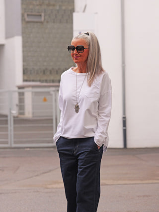 Ältere Frau trägt OFF#DLY LS Fiona white, ein weißes Longsleeve mit Brusttasche, lockerem Schnitt und verlängertem Rücken. Kombiniert mit dunkler Hose und Hamsa-Kette vor heller Wand.