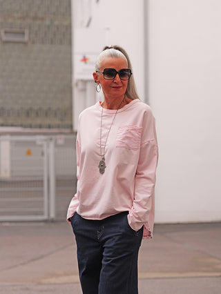Reife Frau trägt OFF#DLY LS Fiona rosa Longsleeve mit lockerer Passform, Brusttasche und langen Ärmeln. Kombiniert mit dunklen Hosen, großen Sonnenbrillen und Hamsa-Kette, zeigt sie stilvolle Eleganz.