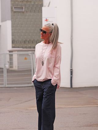 Frau trägt das OFF#DLY LS Fiona rosa Longsleeve mit lockerer Passform, leicht eingesteckt in dunkle, weite Jeans. Sichtbar sind die Brusttasche und der verlängerte Rücken mit Schlitz.