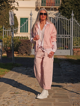 Ältere Frau trägt OFF#DLY Hose Franzie rose: lockerer Schnitt, elastischer Bund, kombiniert mit weiß-hohen Sneakern und rosa Jacke, vor dekorativem Tor posierend.