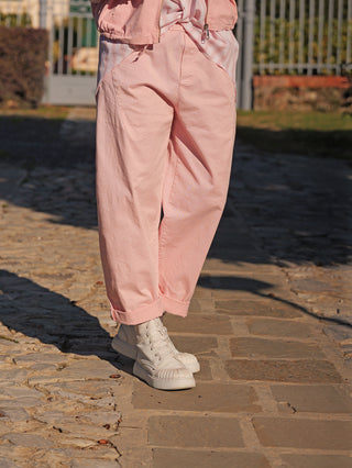 Die OFF#DLY Hose Franzie rose zeigt lässige, pastellrosa Joggpants mit elastischem Bund, lockerem Schnitt und umgeschlagenen Knöcheln, kombiniert mit weißen High-Top-Sneakers auf einem Steinweg.