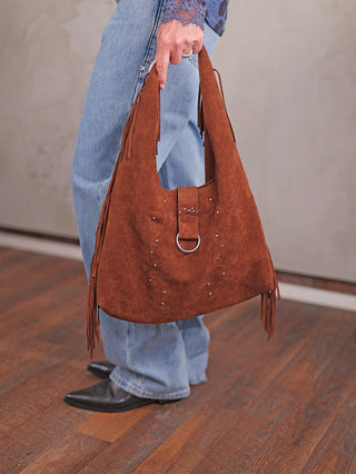 Person hält die OFF#DLY Fransen Bag coffee, eine braune Veloursoptik-Schultertasche mit Fransen, dekorativem Riemen mit silbernem Ring und Nieten, passend zu hellen Jeans und schwarzen Lederschuhen.