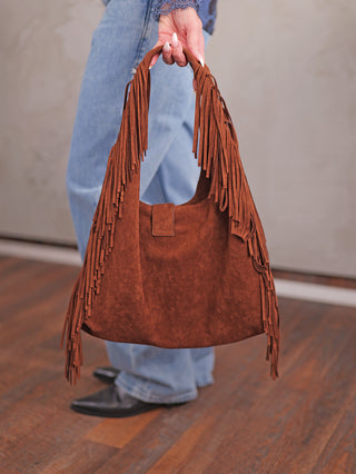 OFF#DLY Fransen Bag coffee: Große, lässige Umhängetasche aus Wildlederimitat in warmem Braun, mit langen Fransen an den Seiten und am Griff, gehalten von einer Person in blauer Jeans und schwarzen Stiefeln.