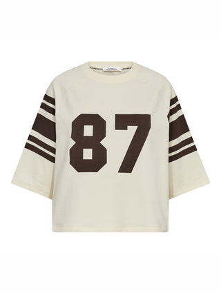 Co Couture Ely 87 Tee Sweat brown/bone zeigt ein cremefarbenes, boxy geschnittenes Shirt mit braunen Varsity-Streifen an den Ärmeln und großer „87“-Aufdruck auf der Brust, verkörpert sportlichen College-Stil.