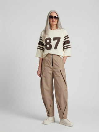 Frau trägt die Co Couture Trice Pleat Pant in Sand, eine locker geschnittene, hoch taillierte Hose mit Falten, elastischem Bund und konischem Bein, kombiniert mit einem sportlichen Oberteil.