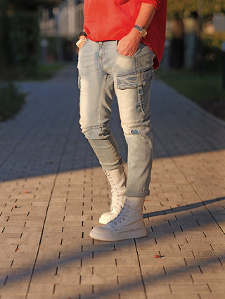 Person trägt OFF#DLY Denim Verona blue: Slim-Fit-Cargo-Jeans mit Flap-Tasche und dezentem Used-Look, kombiniert mit weißen High-Top-Stiefeln. Hände in Taschen gesteckt, Uhr und Armband sichtbar.