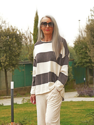 Die Person trägt den OFF#DLY Pulli Ancona black/creme mit breiten Streifen, lockerem Schnitt und langen Ärmeln, kombiniert mit hellen Hosen, in einem Garten stehend.