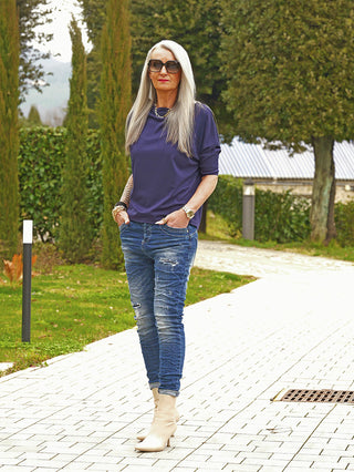 Silberhaarige Frau trägt OFF#DLY Denim Detroy Alassio blue Jeans mit Used-Look und schmaler Passform, kombiniert mit Navy-Top und cremefarbenen Stiefeletten, stehend auf einem Gehweg im Freien.
