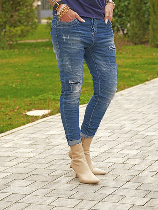 OFF#DLY Denim Detroy Alassio blue Jeans mit schmaler Passform und Destroy-Details, kombiniert mit beigen, spitzen Stiefeletten. Hände mit goldenen Armreifen in den Taschen der lässigen, hochgekrempelten Jeans.