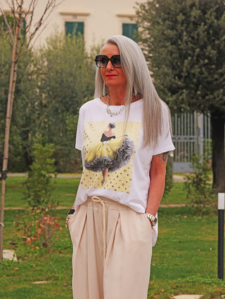 Ältere Frau im Park trägt das OFF#DLY Tee Ballerina white/gelb, mit stilisiertem Ballerina-Print auf weißem T-Shirt, kombiniert mit beigen Hosen und Accessoires.