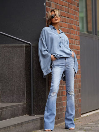 Person lehnt an Gebäudeecke, trägt Gitta Banko Flared Denim Alba Big Sure Blue. Jeans mit leicht ausgestelltem Bein und dezenten Used-Details an den Knien, kombiniert mit lockerem Denim-Hemd.