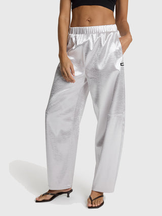 Person trägt ALIX Woven Shiny Pant silver, eine hochgeschnittene, locker sitzende, metallic-silberne Hose mit elastischem Bund und Seitentaschen, kombiniert mit schwarzen Sandalen.