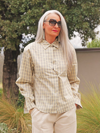 Frau im Freien trägt die OFF#DLY Bluse Lucca yellow Karo, eine langärmelige, pastellkarierte Bluse mit asymmetrischem Überlappungsdetail, kombiniert mit hellbeigen Hosen und stilvollen Accessoires.