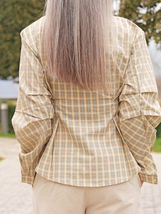 Person von hinten in OFF#DLY Bluse Lucca yellow Karo mit langen Ärmeln und Karomuster, kombiniert mit hellbeigen Hosen, stehend auf einem gepflasterten Weg.