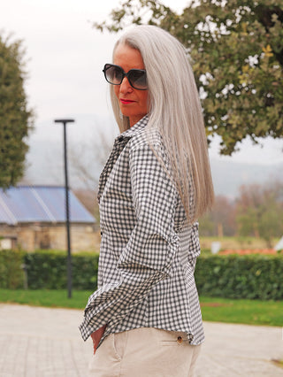 Frau trägt OFF#DLY Bluse Lucca black Karo mit klassischem Karomuster und taillierter Form. Sie schaut über die Schulter in einem Park, gekleidet in helle Hosen und große Sonnenbrille.