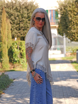 Person mit langen, silber-grauen Haaren trägt das OFF#DLY Tee Shirt Vroni beige, ein fließendes, feminin geschnittenes Oberteil mit Spitzendetails an Ärmeln und Saum, kombiniert mit blauen Jeans.