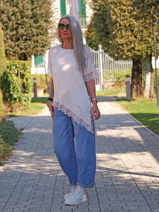 Ältere Frau trägt das OFF#DLY Tee Shirt Vroni rose mit asymmetrischen Spitzenkanten, kombiniert mit blauen Jeans und weißen Sneakers, auf einem Gartenweg stehend.