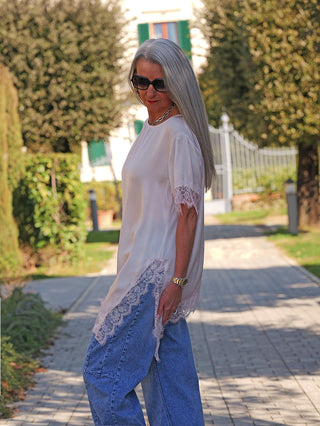 Ältere Frau in oversized Sonnenbrille trägt das OFF#DLY Tee Shirt Vroni rose, ein locker geschnittenes, spitzenverziertes Oberteil, kombiniert mit weiten blauen Jeans, während sie einen Gartenweg entlanggeht.