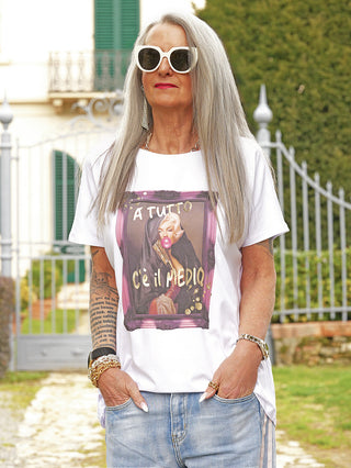 Ältere Frau trägt das OFF#DLY Tee Tutto white mit einem stilisierten Frontprint, kombiniert mit lässigen Jeans, Accessoires und einem Tattoo, vor einem Tor stehend.