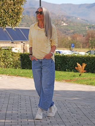 Person trägt den OFF#DLY Ringel Hoody weiß/gelb mit Kapuze und dezentem 3-D Mickey-Maus-Print, kombiniert mit blauer Jeans und weißen Sneakern.