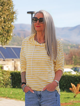 Ältere Frau im OFF#DLY Ringel Hoody Rita, gelb-weiß gestreift, mit Kapuze und fast unsichtbarem Mickey-Maus-Print, in parkähnlicher Umgebung, trägt Jeans und Accessoires.