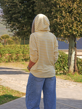 Person trägt den OFF#DLY Ringel Hoody Rita in Gelb-Weiß mit Kapuze, Händen in den Taschen, stehend auf einem Weg, umgeben von Bäumen.