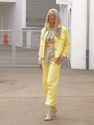 Ältere Frau trägt OFF#DLY Pant Fantasia yellow in Barrel Fit, kombiniert mit einem gelben Ensemble und beigen Stiefeletten, posierend lässig auf urbanem Untergrund.