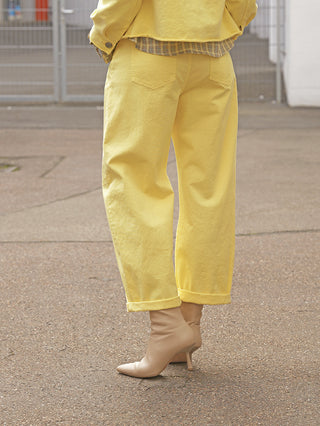 Die OFF#DLY Pant Fantasia yellow zeigt einen modernen Barrel Fit, locker und bequem, aus Baumwoll-Stretch, kombiniert mit spitzen, nudefarbenen Stiefeletten auf einem gepflasterten Untergrund.