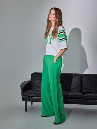 Junge Frau trägt Co Couture Amina Zip Panel Pant in leuchtendem Grün mit schwarzen Streifen und Reißverschlussdetail, kombiniert mit einem weißen, sportlichen T-Shirt, stehend vor schwarzem Ledersofa.