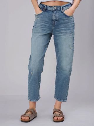 Eine Person trägt True Religion Hope Bow Leg Jeans, leicht verwaschen und leicht zerstört, mit geradem Beinabschluss über den Knöcheln, kombiniert mit taupefarbenen, zweiriemigen Sandalen mit Korksohle.