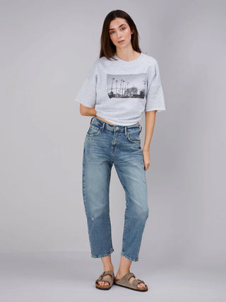 Junge Frau trägt True Religion Hope Bow Leg Jeans Blue, locker geschnitten, schmal zulaufend, mit dezenten Destroyed-Details, kombiniert mit einem grauen T-Shirt mit Palmenmotiv und taupefarbenen Sandalen.