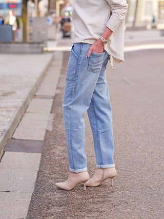 Person trägt die OFF#DLY Joggpant Stella blue mit umgeschlagenem Saum und aufgesetzten Taschen, kombiniert mit nudefarbenen Stiletto-Stiefeletten. Die Jeans zeigt einen lässigen, aber femininen Stil.