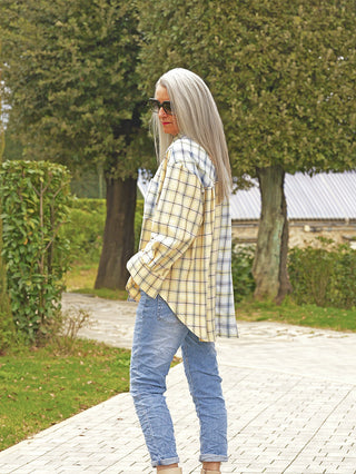 Person mit langen, silbernen Haaren trägt das OFF#DLY Bluse Agliana yellow/blue, ein oversized Karohemd über Jeans, während sie auf einem Gartenweg geht, lässig in Szene gesetzt.