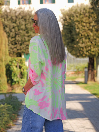 Eine Person trägt die OFF#DLY Bluse Batic green/rose mit leuchtendem Batikmuster und lockerer Passform, kombiniert mit blauen Jeans, in einem sonnigen Gartenweg stehend.