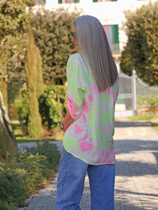 Person von hinten in OFF#DLY Bluse Batic green/rose mit neon-pink und lime-grünem Batik-Muster, lockerem Schnitt und langen krempelbaren Ärmeln, kombiniert mit blauer Relaxed-Fit-Jeans, auf Gartenweg.