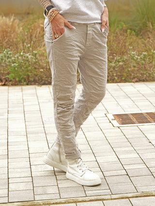 Person trägt OFF#DLY Joggjeans Genua grey, leicht geknitterte Jogger-Hose mit verstellbarem Bund und seitlichen Taschen. Kombiniert mit weißen High-Top-Sneakern und goldenen Armbändern. Hände in den Taschen.