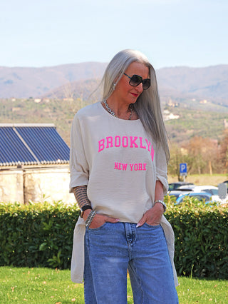 Erwachsene Frau in Garten mit OFF#DLY Longshirt Tamara nature, beige mit neonpinken „Brooklyn New York“-Print. Lockerer Schnitt, lässig zu Jeans. Accessoires und Tattoos ergänzen den Look.