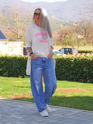 Eine Frau trägt das OFF#DLY Longshirt Tamara nature mit „BROOKLYN“-Schriftzug, kombiniert mit weiten Jeans und Sneakers, während sie in einem parkähnlichen Umfeld steht.