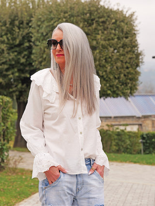 Frau trägt die OFF#DLY Bluse Greta white mit Spitzenkragen und -ärmelabschlüssen. Sie kombiniert die Bluse mit lässigen Jeans, stehend auf einem gepflasterten Weg in einem Garten.