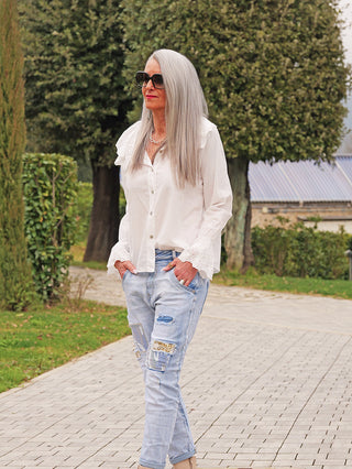 Person trägt die OFF#DLY Bluse Greta white mit großzügigem Spitzenkragen und Knopfleiste, kombiniert mit Patchwork-Jeans, während sie auf einem Gartenweg spazieren geht.