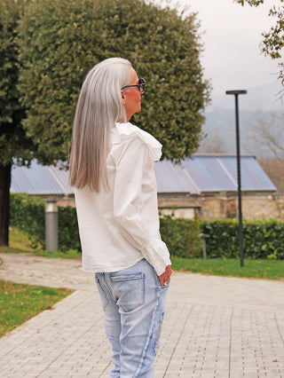 Person in Park trägt die OFF#DLY Bluse Greta white mit großem Spitzenkragen und Spitzen-Ärmelabschlüssen, kombiniert mit hellen, leicht zerrissenen Jeans für einen eleganten, femininen Look.